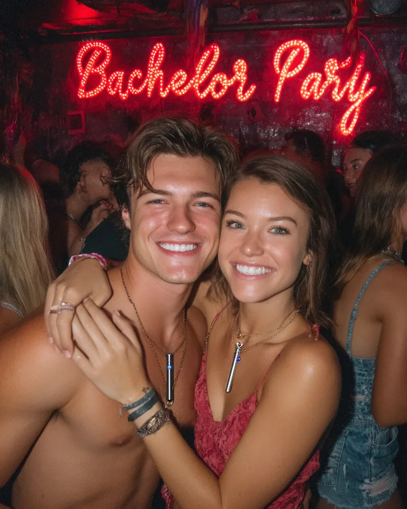 A man and woman embracing at a singles party, both wearing the Lumea bullet vibrator necklace, enjoying discreet pleasure together